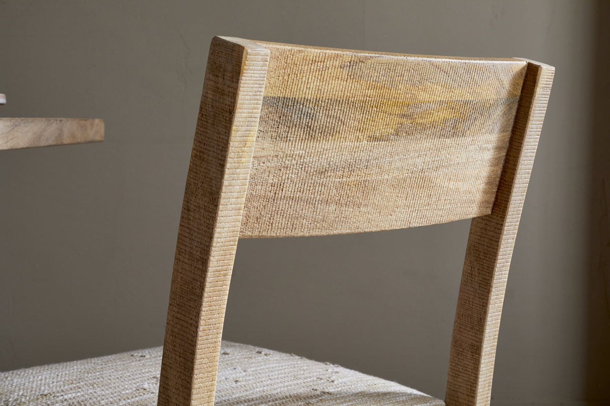 Close-up of the nkuku Zoha Cotton & Jute Upholstered Dining Chair in Natural, featuring a flat, angled backrest, visible wood grain, and a seat covered in light cotton and jute fabric. Shown next to a matching table.