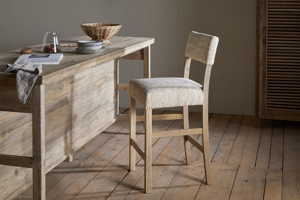 A rustic kitchen features the nkuku Zoha Cotton & Jute Upholstered Counter Chair in natural, set at a wooden table styled with a towel, glass, pitcher, book, plates, and wicker basket amid neutral minimalist décor.