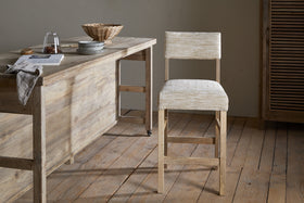 A rustic mango wood kitchen features the nkuku Zoha Cotton & Jute Upholstered Counter Chair in Natural, with a textured cushion. A wooden table displays cookies, a glass jar, woven basket, bowl, and open book on light wood floors and neutral walls.