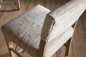 A close-up of the nkuku Zoha Cotton & Jute Upholstered Counter Chair in Natural shows its textured cream seat and backrest, with sunlight highlighting the fabric and stitching. Another Zoha chair is partially visible in the background.