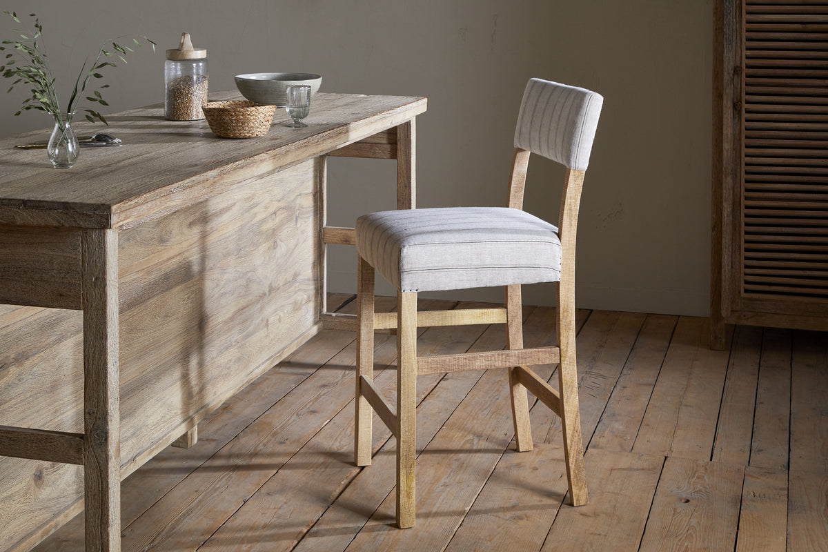 The Nidhi Linen Upholstered Counter Chair by nkuku in natural mango wood sits beside a matching table holding a vase with a branch, jar, bowl, and cup. Wooden floors and neutral walls complete the cozy minimalist vibe.