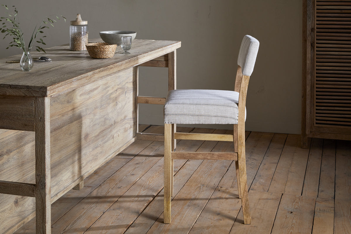 The Nidhi Linen Upholstered Counter Chair by nkuku, with its padded light beige seat, sits beside a rustic mango wood dining table accented with greenery and ceramics. The space features earthy tones and a wooden floor.