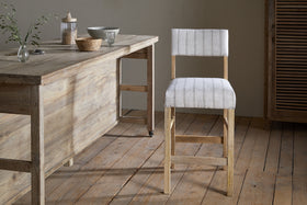 The Nidhi Linen Upholstered Counter Chair by nkuku features striped white linen on a mango wood frame. It sits on a wooden floor beside a rustic table topped with greenery, baskets, and a lidded jar in a softly lit, neutral-toned room.