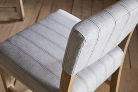 A close-up, angled view of the nkuku Nidhi Linen Upholstered Counter Chair - Natural, featuring light gray linen with thin vertical stripes and a mango wood frame, set on a light brown wooden floor. Another chair appears slightly out of focus in the background.
