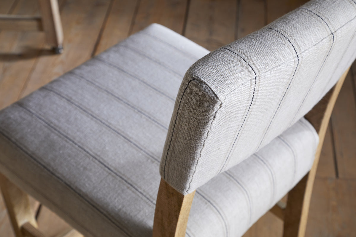 A close-up, angled view of the nkuku Nidhi Linen Upholstered Counter Chair - Natural, featuring light gray linen with thin vertical stripes and a mango wood frame, set on a light brown wooden floor. Another chair appears slightly out of focus in the background.