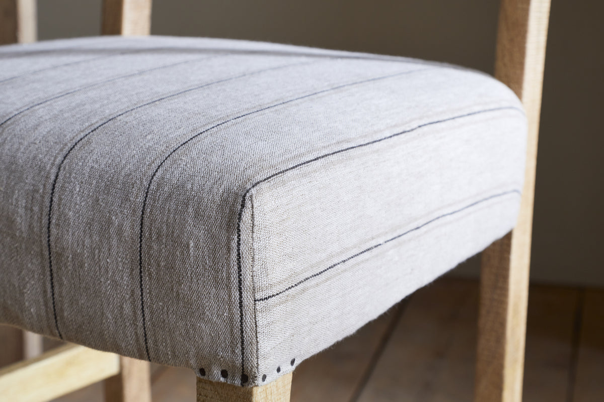 A close-up of the Nidhi Linen Upholstered Counter Chair - Natural by nkuku shows mango wood, a light gray linen seat with dark stripes, textured fabric, and small decorative nailheads at the corners.