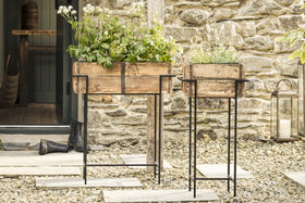 Two rustic reclaimed wooden planter boxes with green and white plants rest on tall nkuku Amazzi Indoor Planter Stands in Natural & Black, placed on a pebble patio before a stone wall. A door, black boot, and lantern enhance the cozy outdoor scene.