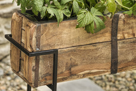 The nkuku Amazzi Indoor Planter Stand in Natural & Black features reclaimed wood with visible grain and black metal accents, perfect for showcasing indoor plants. Pebbles and a stone surface appear softly blurred in the background.