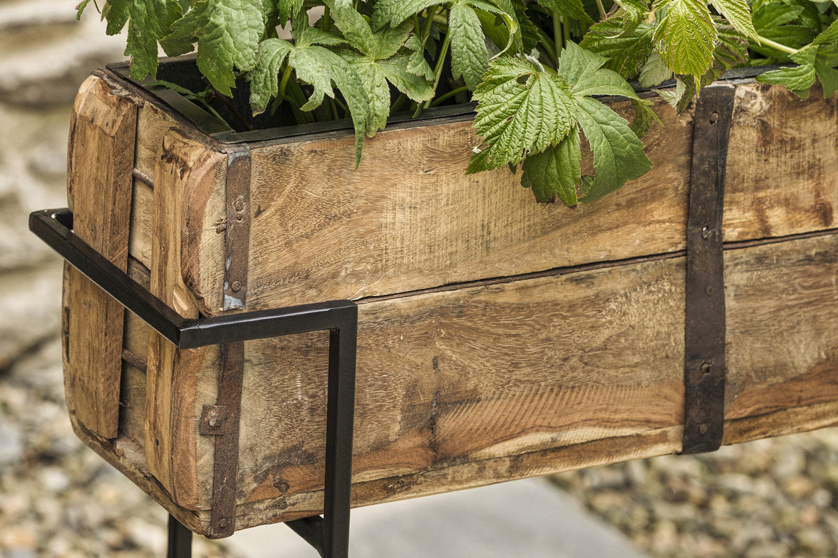The nkuku Amazzi Indoor Planter Stand in Natural & Black features reclaimed wood with visible grain and black metal accents, perfect for showcasing indoor plants. Pebbles and a stone surface appear softly blurred in the background.