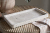 The nkuku Ganda Travertine Vanity Tray - Cream, featuring a smooth, slightly textured rectangular surface, rests on a round wooden table with carved grooves, paired with a brown-and-white striped vase and a folded beige and white cloth.