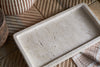 The nkuku Ganda Travertine Vanity Tray in Cream, featuring a rough, porous finish, rests on a wooden geometric surface beside a striped beige cloth, round wooden container, and woven basket for a neutral bathroom organiser look.