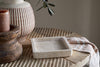The Ganda Travertine Vanity Tray - Cream by nkuku sits on a textured wooden table, with a striped ceramic vase, a rustic brown pillar candle, and a striped cloth nearby. Olive leaves hang above, adding natural elegance to the scene.