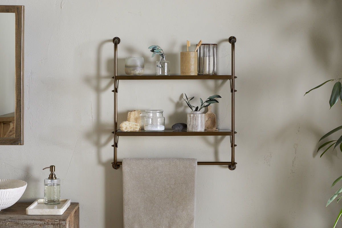 The Karura Wall Shelves with Towel Rail in Antique Brass by nkuku hold jars, a toothbrush holder, cups, a loofah, stones, and small plant cuttings. A beige towel hangs below; to the left sit a mirror and soap dispenser on a wooden counter.