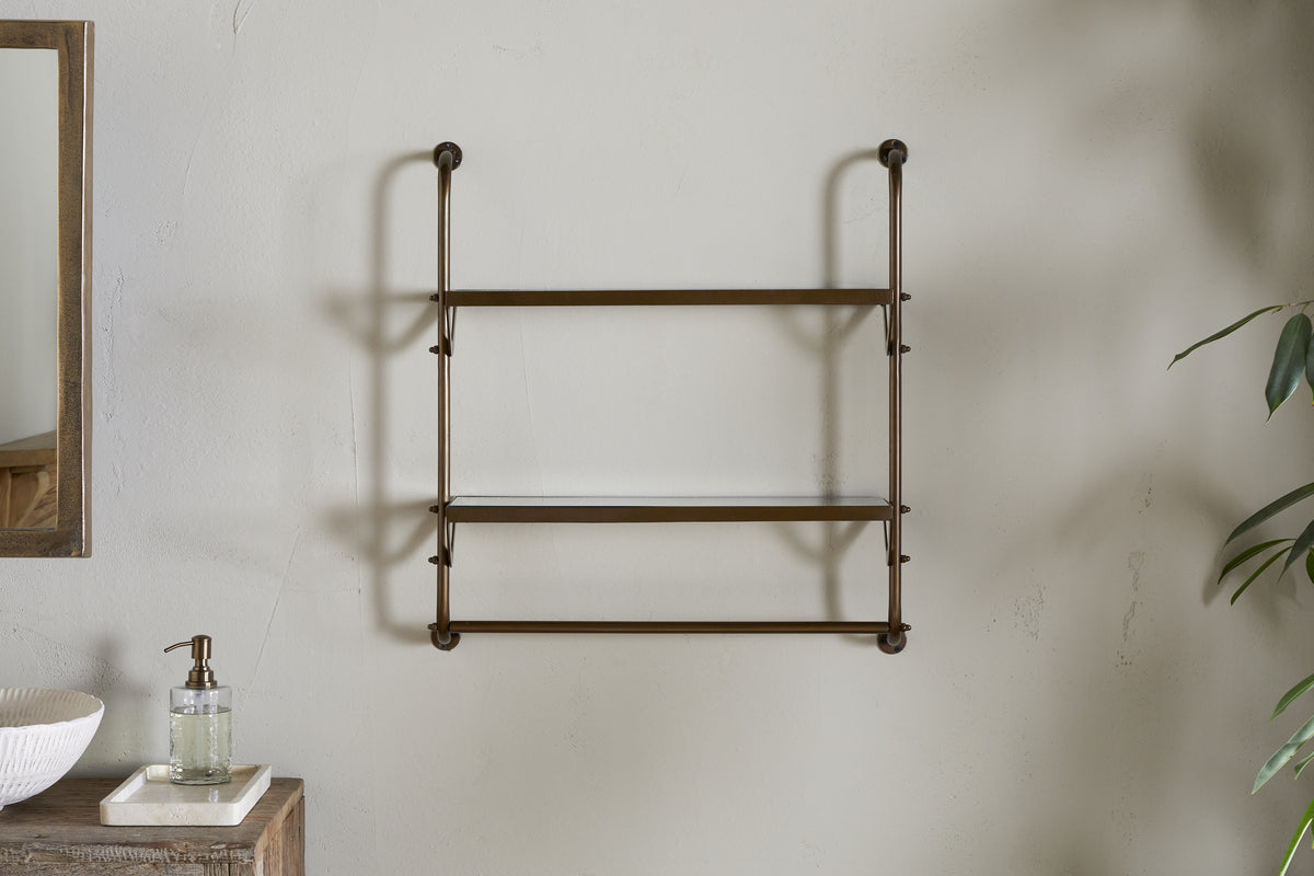 The nkuku Karura Wall Shelves with Towel Rail in Antique Brass, featuring two glass shelves, is mounted on a light gray wall. Part of a mirror, a tray with soap dispenser, and green plant leaves are also visible in the image.