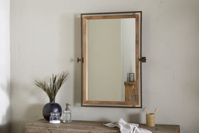 The nkuku Mar Swivel Wall Mirror in Antique Brass & Natural is mounted on a light beige wall above a rustic wooden table, which holds a black vase with green stems, clear soap dispenser, bamboo toothbrush cup, and folded white cloth.