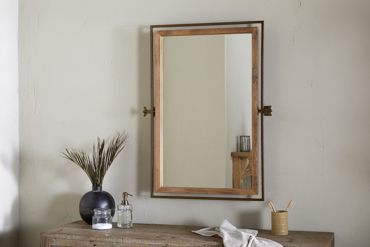 The nkuku Mar Swivel Wall Mirror in Antique Brass & Natural is mounted on a light beige wall above a rustic wooden table, which holds a black vase with green stems, clear soap dispenser, bamboo toothbrush cup, and folded white cloth.
