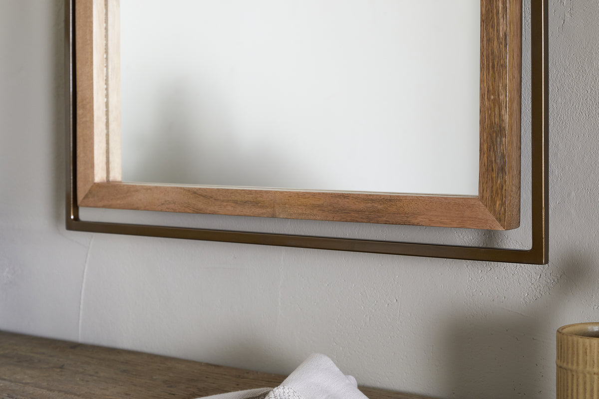 A close-up of the nkuku Mar Swivel Wall Mirror in Antique Brass & Natural, featuring a rectangular mango wood frame, mounted on a light wall. Below it, a wooden surface holds a white cloth and beige ribbed container.