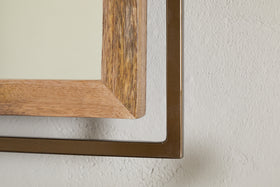 Close-up of the Mar Swivel Wall Mirror by nkuku, featuring natural mango wood and an antique brass metal bracket at the corner, mounted on a textured off-white wall. Only the detailed corner is visible, highlighting its industrial style.