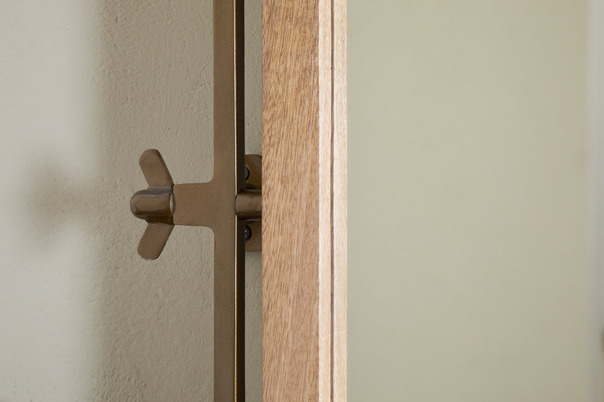 A close-up of the nkuku Mar Swivel Wall Mirror - Antique Brass & Natural shows its brown metal bracket with a wingnut attached, mounted on a light wall. The antique brass edge and visible wood grain highlight its elegant design.