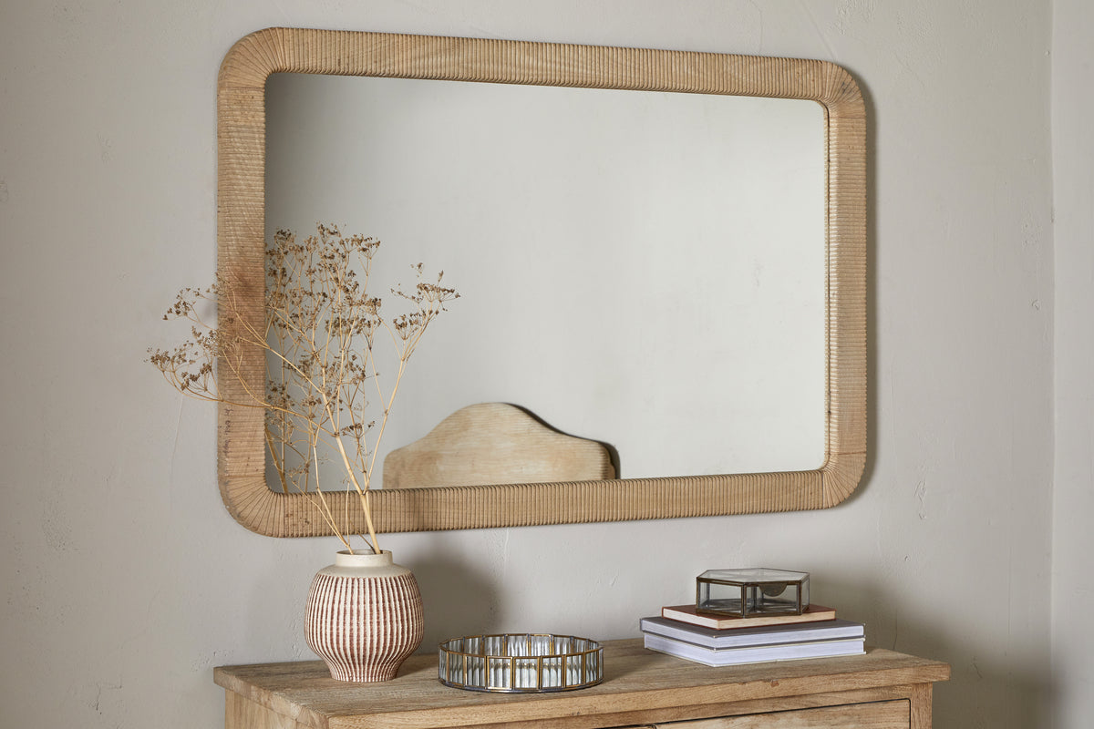 The nkuku Vivan Rectangular Grooved Wood Mirror in natural finish hangs above a wooden dresser topped with a striped vase of dried flowers, round tray, glass box, and books, set against a plain light-colored wall.