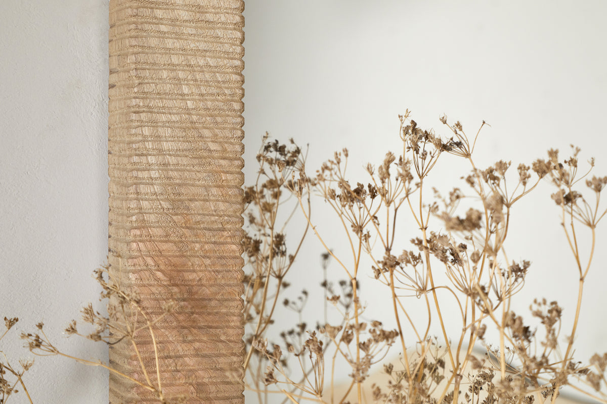 A close-up of dried beige wildflowers with slender stems beside the nkuku Vivan Rectangular Grooved Wood Mirror - Natural. The light, plain background highlights the mirror's vertical ribbed mango wood frame and minimalist design.