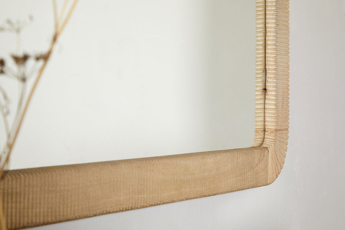 A close-up of the nkuku Vivan Rectangular Grooved Wood Mirror in natural, featuring a hand-carved mango wood frame with textured grooves, mounted on a white wall. Soft-focus dried stems appear on the left.