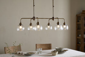A dining room features a light wood table set for a meal with plates, bowls, and glasses. Overhead, the nkuku Kakira Chandelier Pendant in Antique Brass & Clear casts a warm glow. Wooden chairs, a sprig in glass, and a wooden cabinet complete the scene.