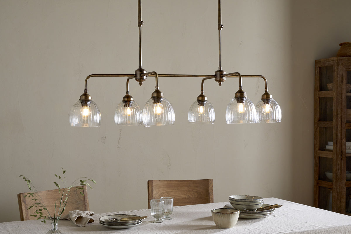 A dining room features a light wood table set for a meal with plates, bowls, and glasses. Overhead, the nkuku Kakira Chandelier Pendant in Antique Brass & Clear casts a warm glow. Wooden chairs, a sprig in glass, and a wooden cabinet complete the scene.