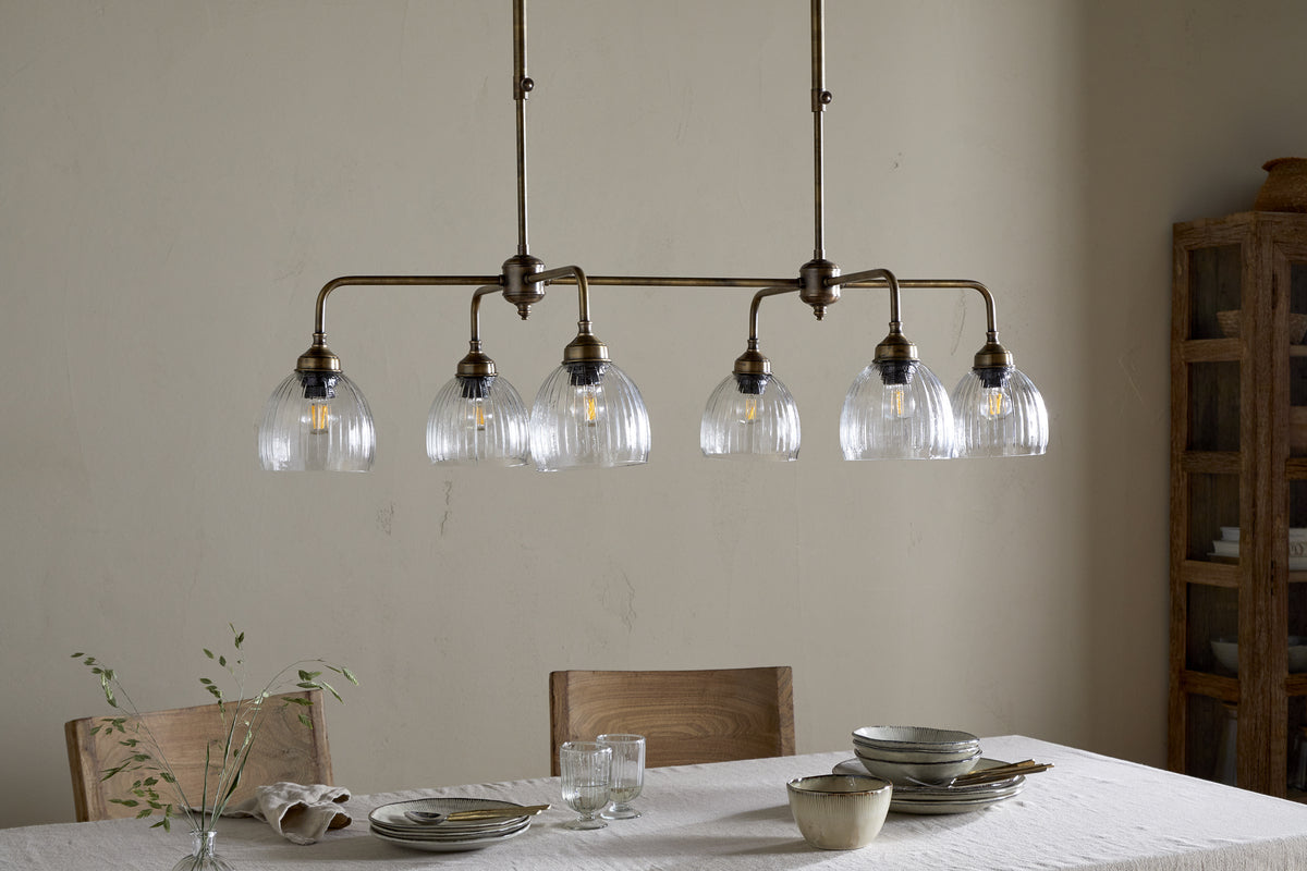 A dining table set with plates, bowls, glasses, and utensils sits below the nkuku Kakira Chandelier Pendant in antique brass with five ribbed glass shades. Wooden chairs and a cabinet complete the softly lit room.