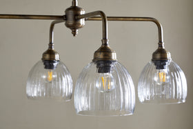 The Kakira Chandelier Pendant by nkuku features antique brass and clear ribbed bell-shaped glass shades. Three exposed bulbs hang from a horizontal rod, creating a vintage-industrial focal point for any space.