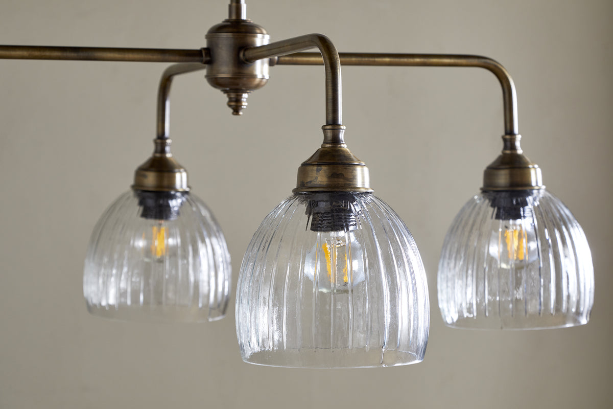 The Kakira Chandelier Pendant by nkuku features antique brass and clear ribbed bell-shaped glass shades. Three exposed bulbs hang from a horizontal rod, creating a vintage-industrial focal point for any space.