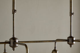 Close-up of the nkuku Kakira Chandelier Pendant in Antique Brass & Clear, featuring visible rods and joints, clean lines, metal hardware, and a muted finish—all set against a plain beige wall for an industrial yet elegant look.
