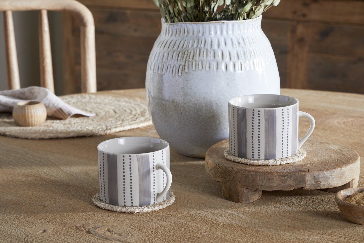 Two large cream and grey Patthar Mugs by nkuku rest on woven coasters atop a rustic wooden table, one elevated on a small stand. A white textured vase with dried flowers is in the background, near a chair and placemat.