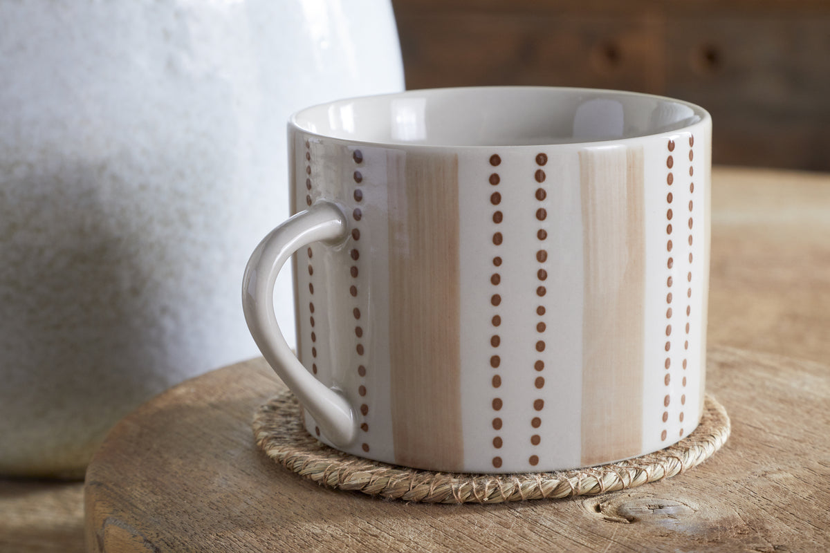 The Patthar Mug - Cream & Clay - Large (Set of 2) by nkuku, featuring beige and white stripes and dotted lines, rests on a woven coaster atop wood. A large white vase in the background adds to the cozy, rustic vibe.