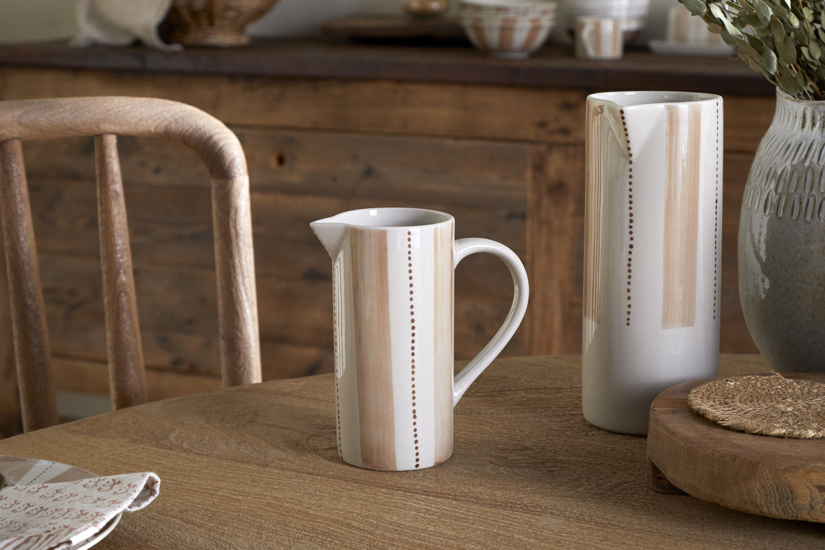 The nkuku Patthar Jug - Cream & Clay - Small, with hand-painted vertical beige stripes and dotted accents, sits on a wooden table beside a matching vase and rustic decor elements—an inviting touch for any tableware collection.