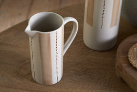The nkuku Patthar Jug - Cream & Clay - Small, hand-painted with vertical tan stripes and dotted lines, sits on a wooden table beside its matching tall jug, a woven mat, and a round tray for a cozy rustic look.