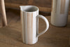 The nkuku Patthar Jug - Cream & Clay - Small, with a tall cylindrical shape, curved handle, and hand-painted tan stripes with dotted lines, sits on a wooden table. A matching jug adds to the warm, rustic background.