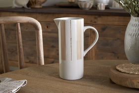 The nkuku Patthar Jug - Cream & Clay - Large, a tall hand-painted ceramic piece with delicate vertical dotted lines, sits on a wooden table set with a napkin, woven placemat, and large vase. A rustic chair and sideboard complete the cozy scene.