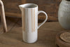 The nkuku Patthar Jug – Cream & Clay – Large, a tall hand-painted ceramic piece with a curved handle in white with beige stripes and dots, sits on a wooden table. A textured vase and wooden chair complete the rustic, cozy scene.