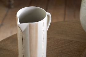 The Patthar Jug - Cream & Clay - Large by nkuku, featuring hand-painted vertical beige stripes and dotted patterns on white ceramic, stands on a round wooden table with soft natural light and a blurred wooden floor in the background.