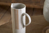 The Patthar Jug - Cream & Clay - Large by nkuku, a tall white ceramic jug with a curved handle, features hand-painted dotted vertical lines and a beige stripe. It sits on wood, with its spout visible and another textured ceramic item partly in view.