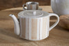 The Patthar Teapot - Cream & Clay - Large by nkuku sits on a wooden table, hand-painted with cream tones, beige stripes, and brown-dot lines. A matching cup and saucer from nkuku appear blurred in the background.