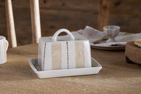 The Patthar Butter Dish - Cream & Clay by nkuku, hand-painted with vertical beige stripes and dotted lines, sits on a wooden table. Dishes, a napkin, and a wooden chair complete the cozy, rustic kitchen scene.