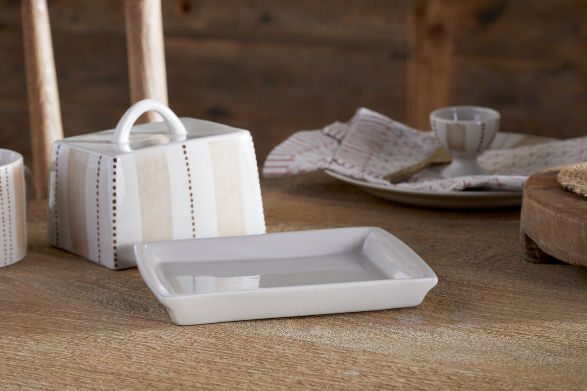 The Patthar Butter Dish - Cream & Clay by nkuku sits on a wooden table, its hand-painted lid and handle adding rustic charm. Nearby are a plate with napkins and a candle holder, perfect for cozy decorative kitchen storage.