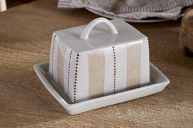 The nkuku Patthar Butter Dish - Cream & Clay is a hand-painted ceramic piece with a looped handle, featuring vertical beige stripes and dotted brown lines. It sits on a wooden table, with kitchen storage plates stacked in the background.