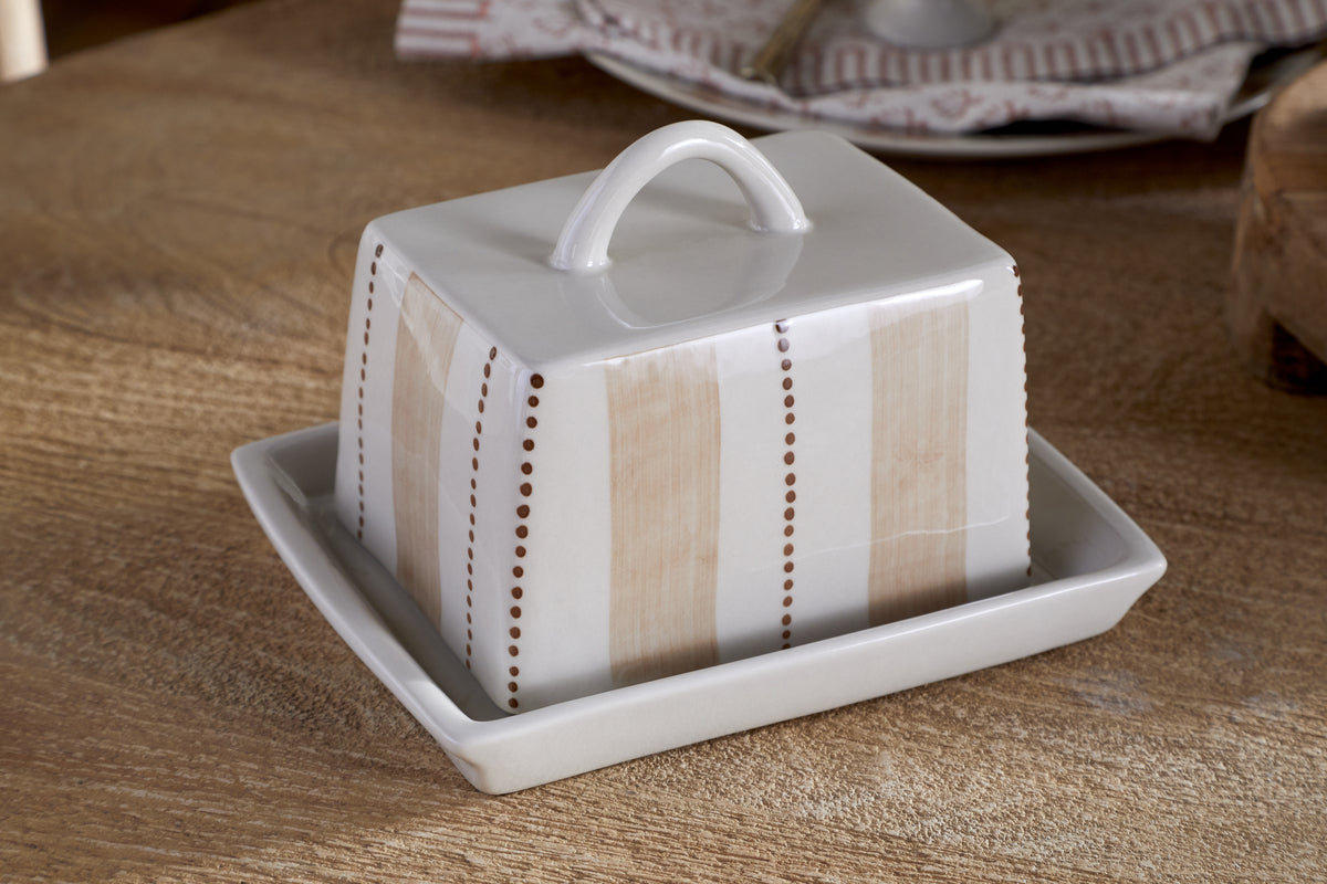 The nkuku Patthar Butter Dish - Cream & Clay is a hand-painted ceramic piece with a looped handle, featuring vertical beige stripes and dotted brown lines. It sits on a wooden table, with kitchen storage plates stacked in the background.