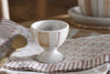 The nkuku Patthar Egg Cups – Cream & Clay (Set of 2) feature white ceramic with beige stripes and dots, shown on a patterned napkin with a gold spoon. Patthar dinnerware in the background adds rustic charm to the table setting.