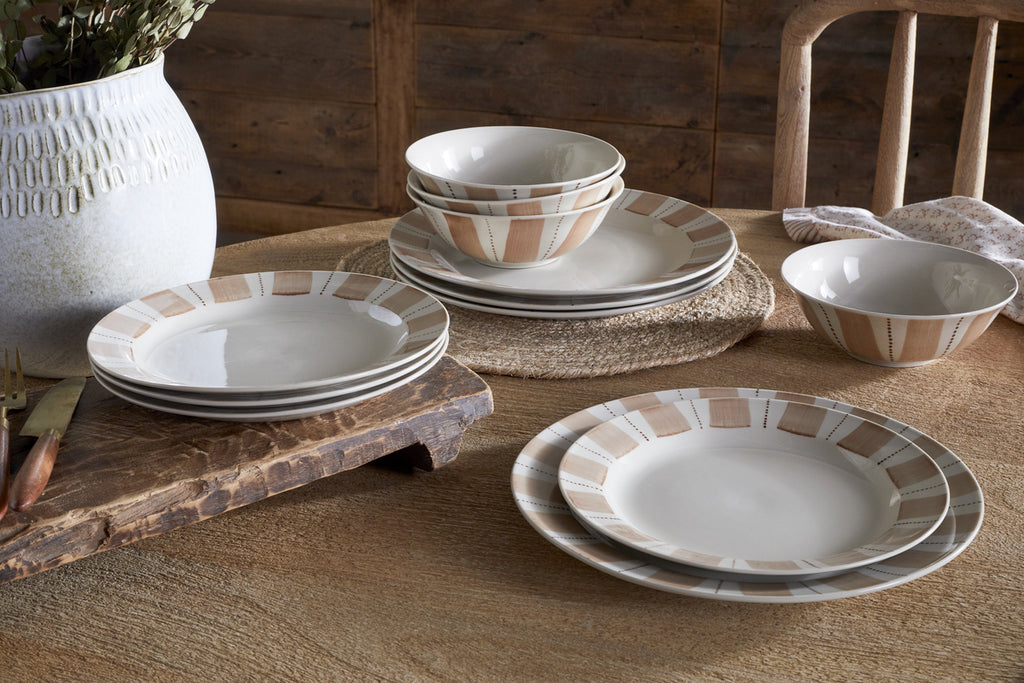 A rustic wooden table is set with nkuku’s Patthar Dinnerware Set - Cream & Clay (set of 12), some dishes stacked, others arranged. A textured white vase with dried flowers and a wooden cutting board add to the cozy dining scene.