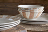 A close-up of the nkuku Patthar Dinnerware Set - Cream & Clay (Set of 12) showcases hand-painted beige and brown patterns, stacked on a woven placemat atop wood for a rustic, cozy vibe.