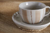 The nkuku Patthar Cup & Saucer - Cream & Clay, featuring vertical beige and brown dotted stripes, rests on a woven coaster atop a light wooden table, softly lit to highlight its charming handmade appeal.